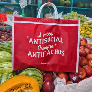 Bolsa roja de mercado con asas blancas y el mensaje tipográfico "A veces ANTISOCIAL Siempre ANTIFACHOS", colocada frente a un puesto de frutas.